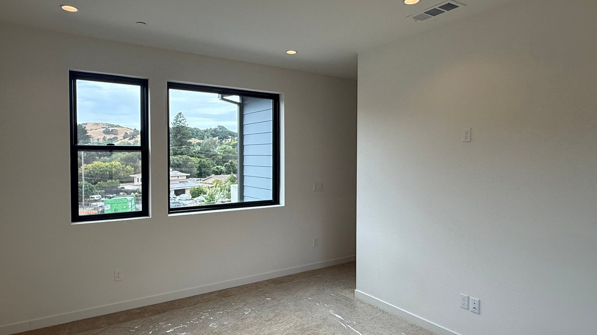 Empty room with white walls, two windows, and a view of greenery outside.