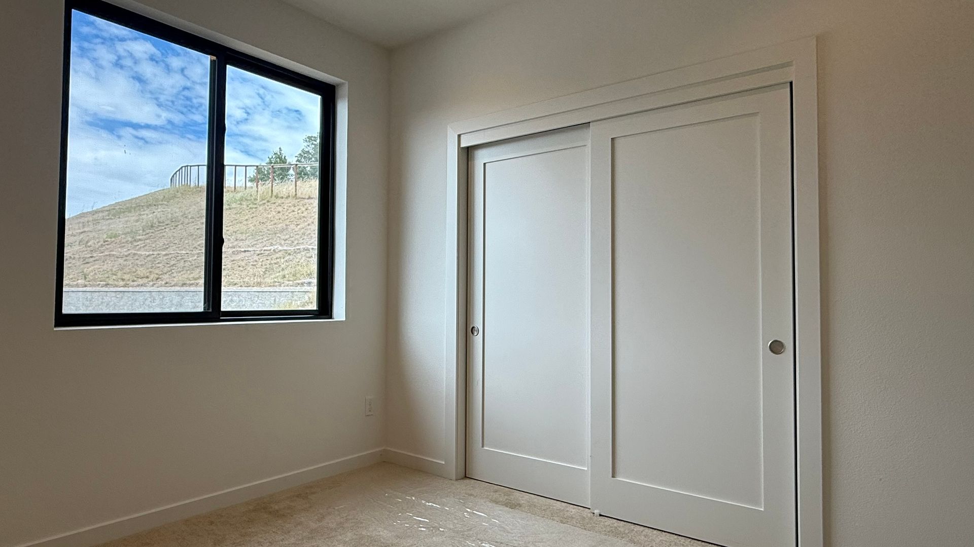 Empty beige bedroom with a black-framed window and white closet doors