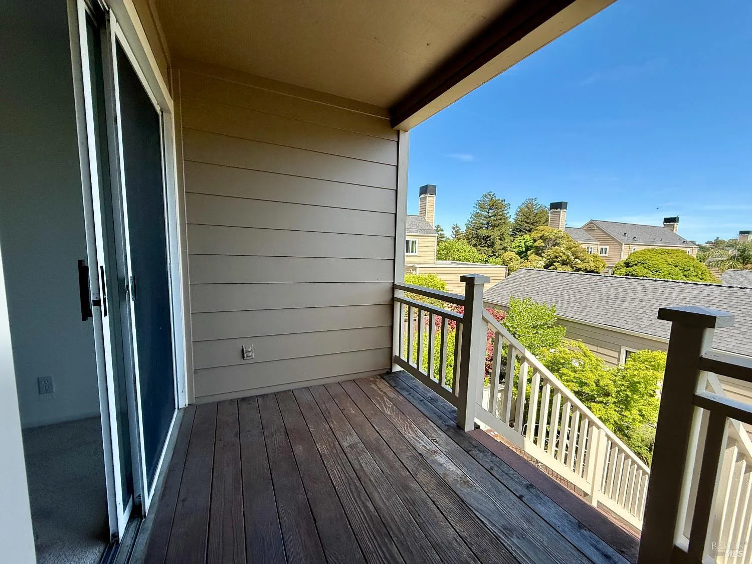 Covered apartment balcony with wood floor, sliding glass door, and railing overlooking rooftops and trees