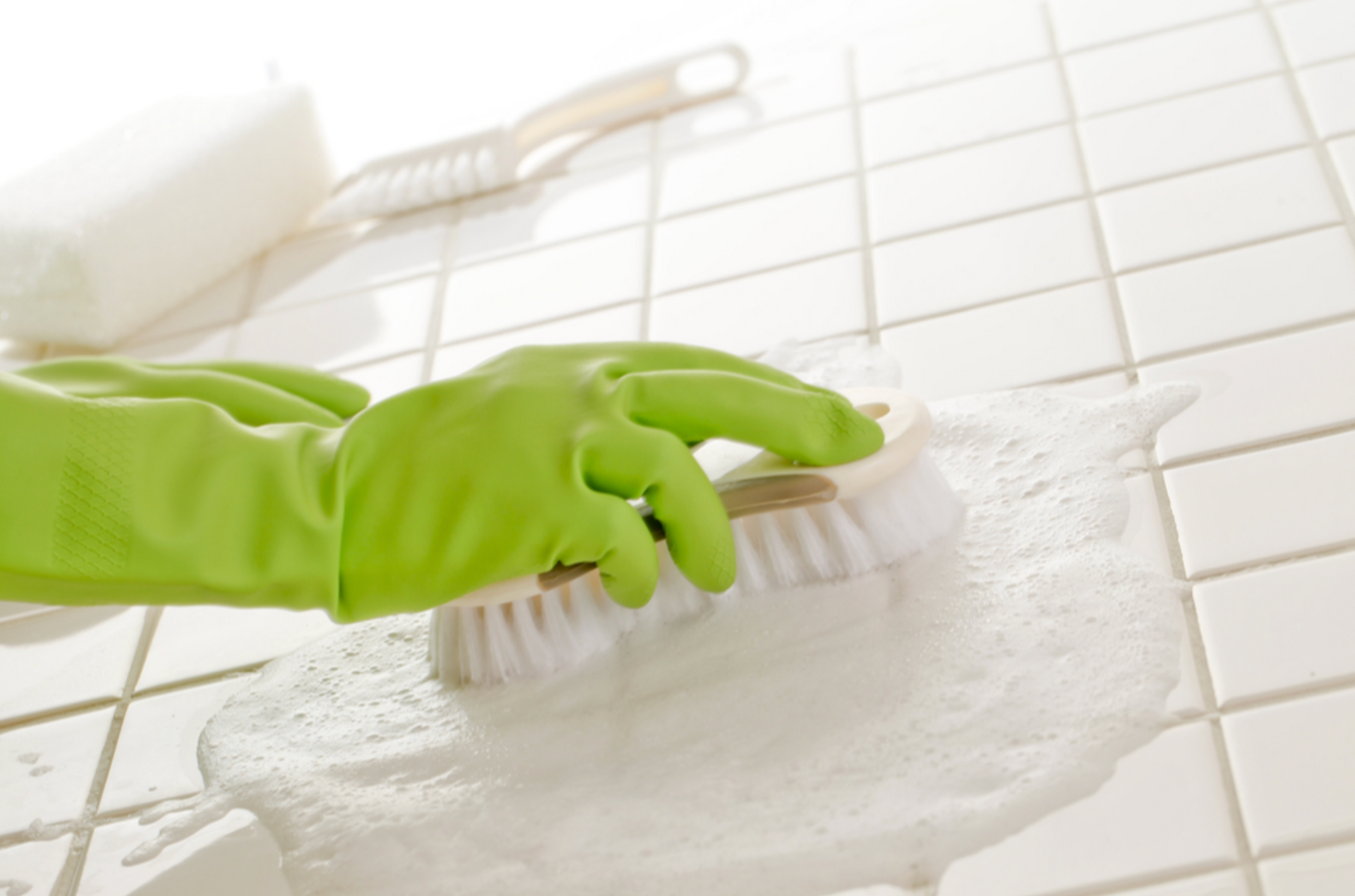 Green rubber gloves using a scrub brush and soap suds to clean white bathroom tiles. 