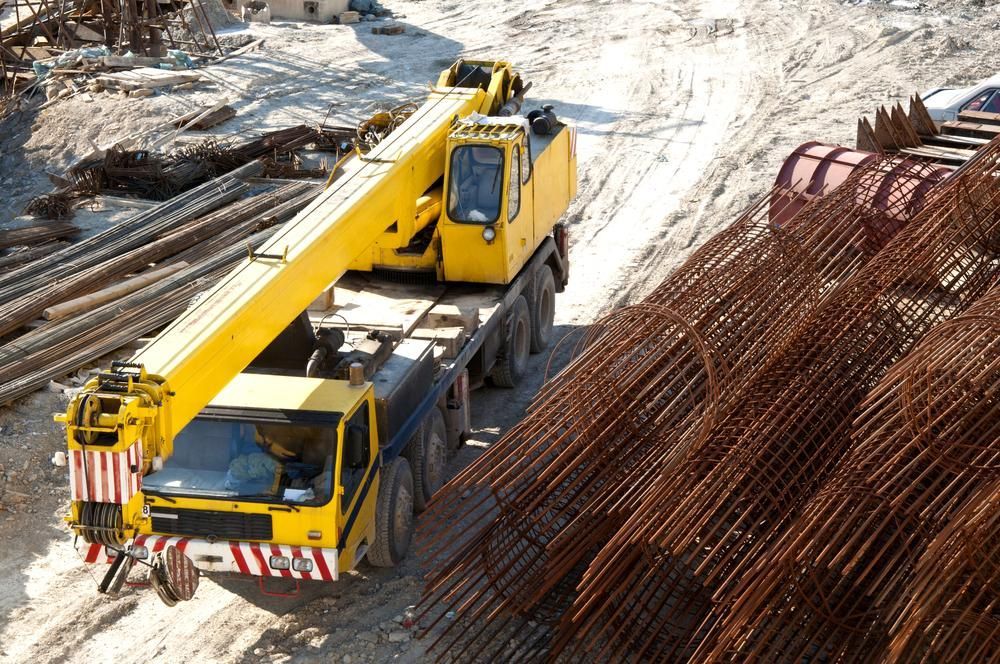 A Yellow Crane Is Driving Down a Dirt Road Next to A Pile of Steel Bars — AMAC Cranes in Coolum Beach, QLD