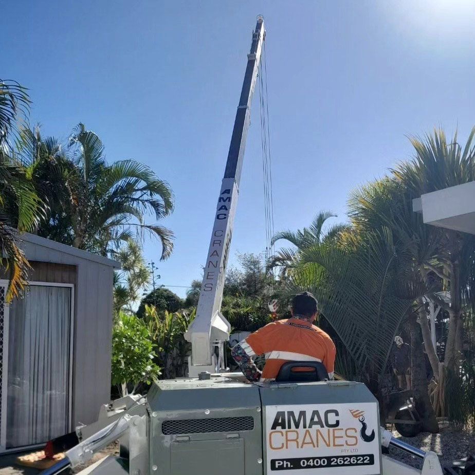 A Mobile Crane Operated by a Person in an Orange Vest, Lifting Something Near a House and Palm Trees Under a Blue Sky