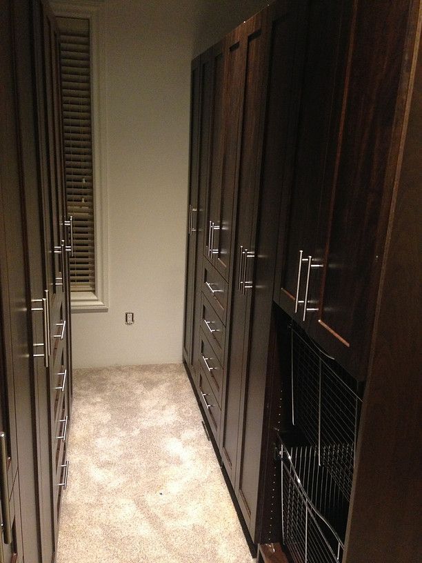 Narrow walk-in closet with dark wood cabinets and hanging storage on both sides, beige carpet, and a window at the end