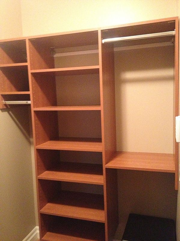Open wooden closet with shelves, hanging rods, and a lower cubby in a beige room.