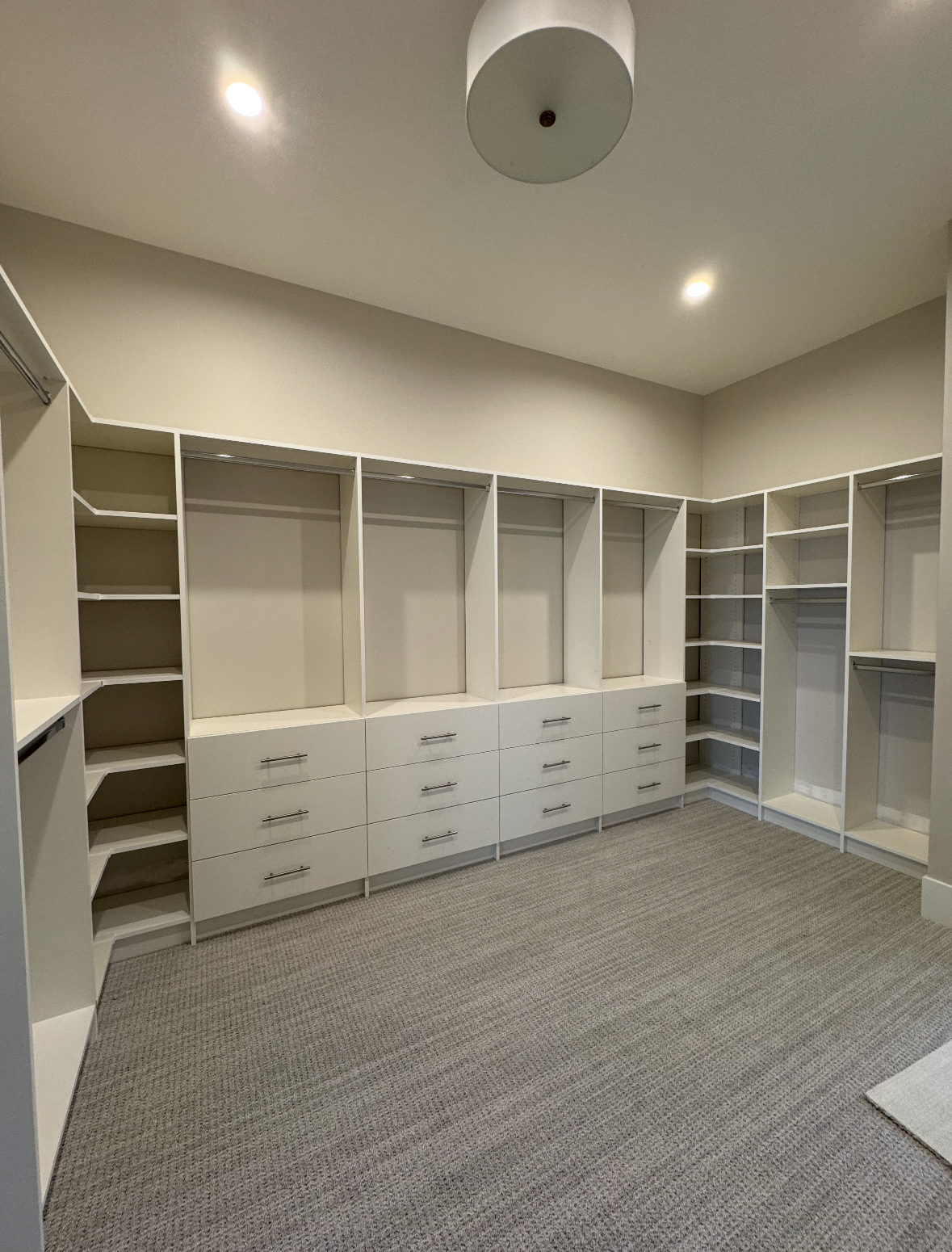 Empty walk-in closet with white built-in shelves, drawers, and gray carpet under recessed ceiling lights