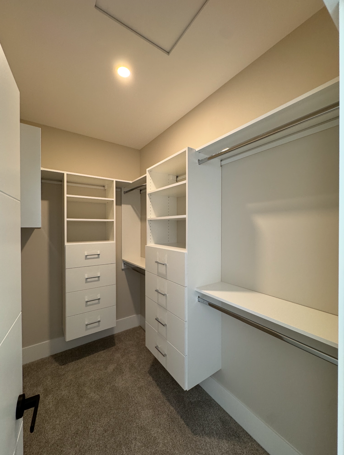 Empty walk-in closet with white shelving, drawers, and hanging rods on beige walls and carpet.