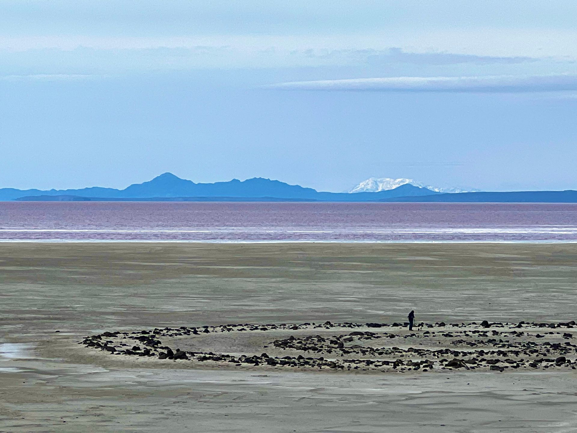 Person stands in a stone spiral on a salt flat, pink water, distant mountains, and pale blue sky.