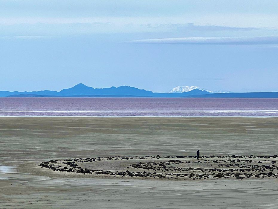 Person stands in stone circle on a sandy, muddy shore with pink water and mountains under a blue sky.