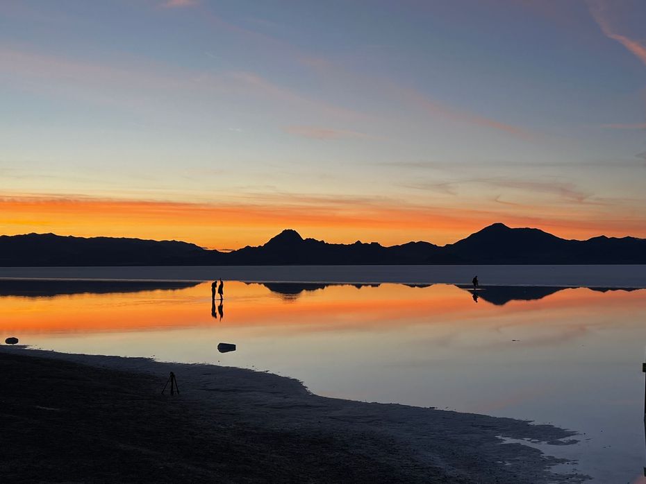 Sunset over calm water with silhouettes of mountains and people, reflecting fiery orange and blue sky.