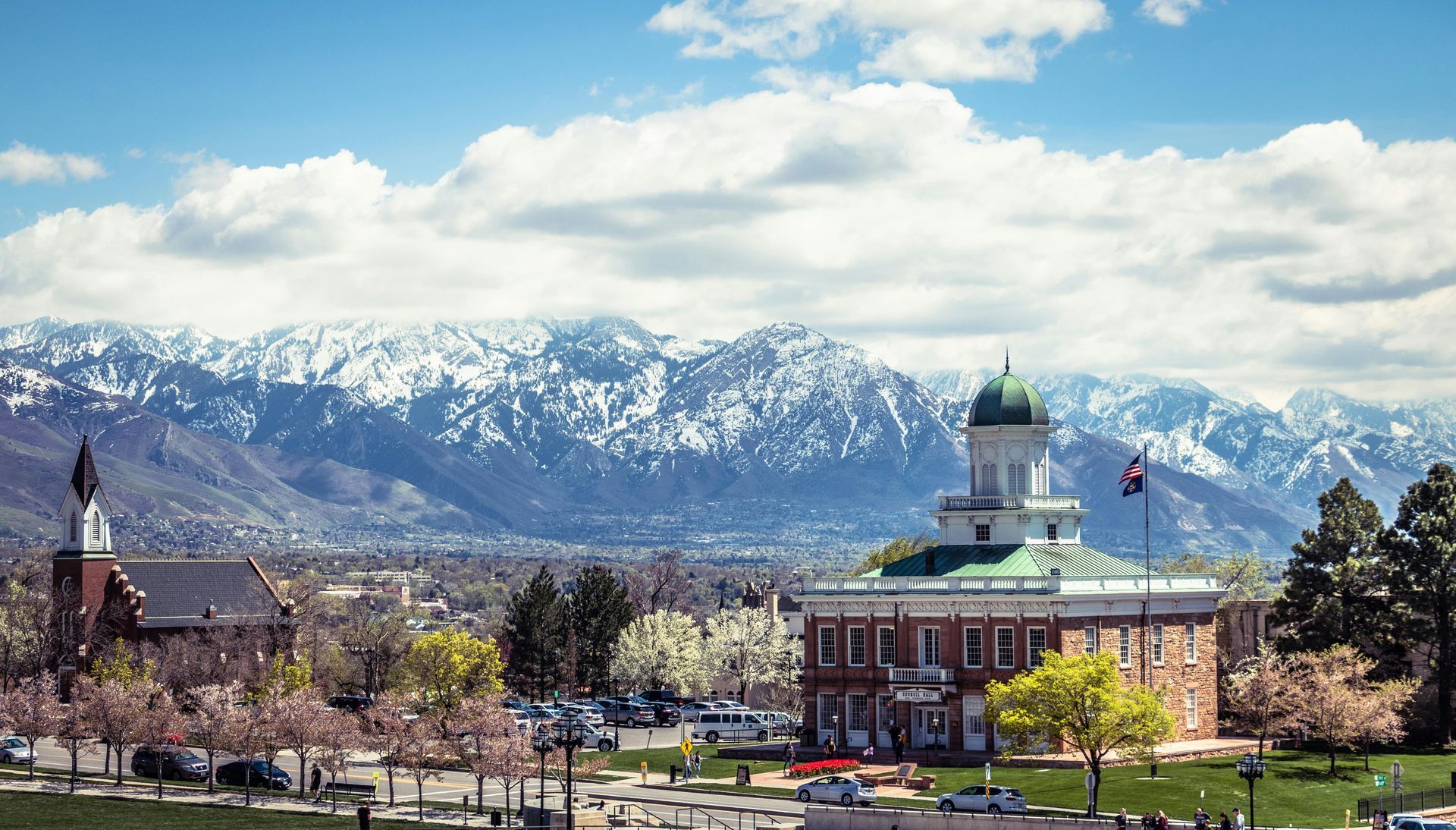 Historic building with green dome, trees, and snow-capped mountains under a blue sky.