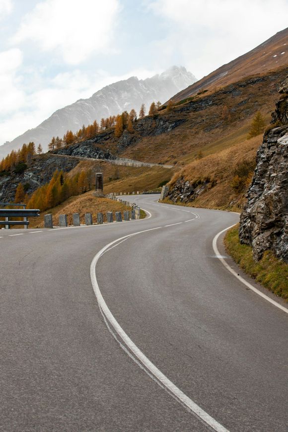 Curving mountain road with yellow trees, rocky cliffs, and a snow-capped peak in the distance.