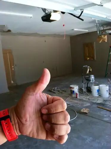 Hand giving a thumbs up in a partially renovated garage. Paint supplies visible.