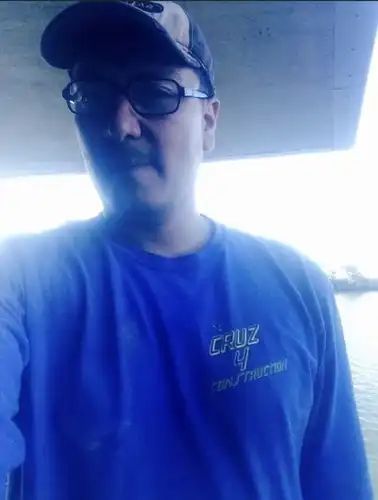 Man in blue shirt, baseball cap, and glasses under a structure, looking at the camera.