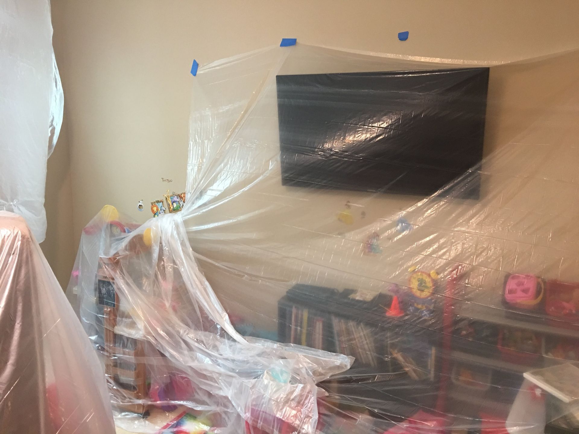 Clear plastic sheeting covers a wall-mounted TV and children's furniture in a room. Blue tape secures the plastic.