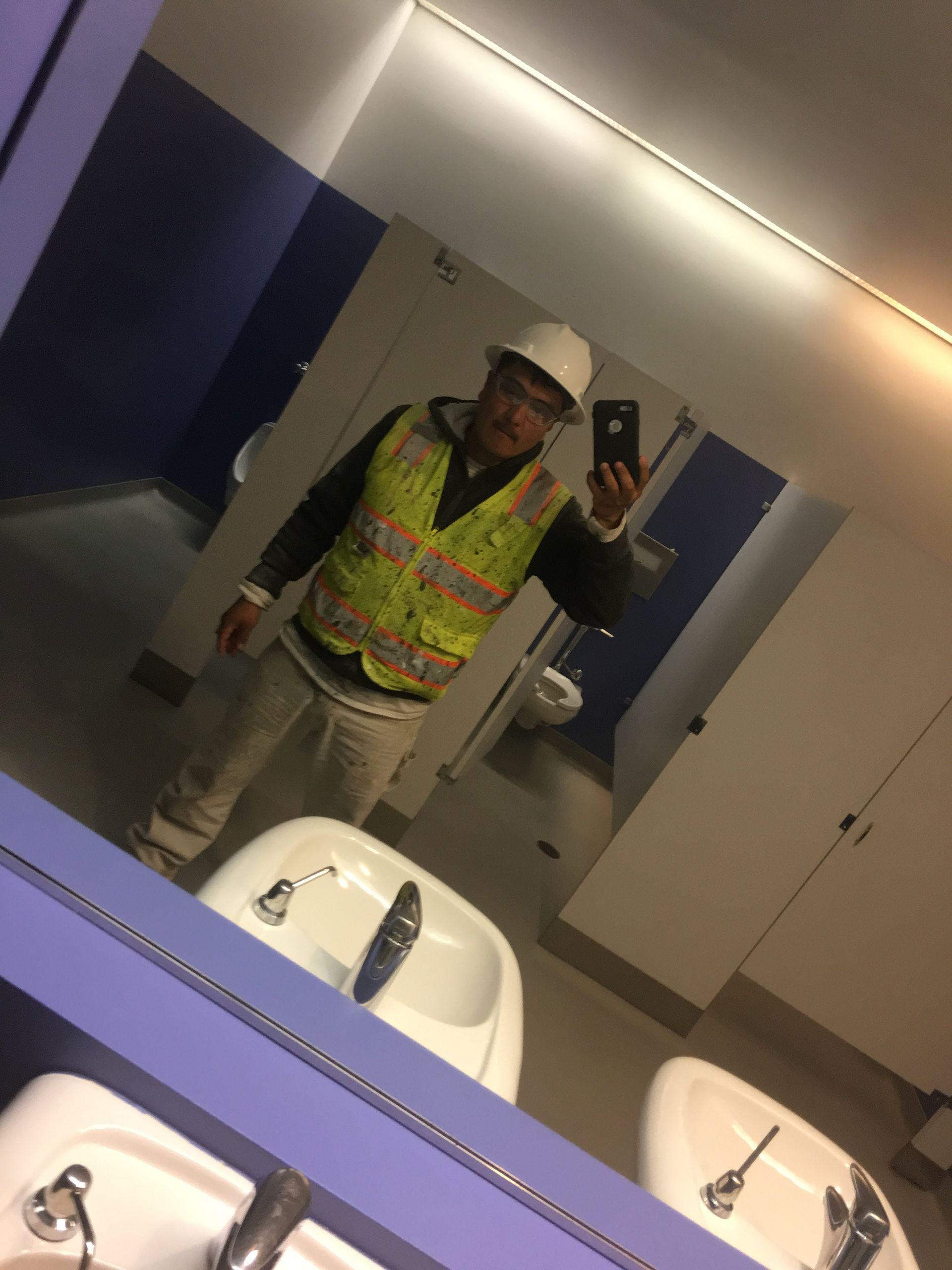 Person in construction attire taking a selfie in a public restroom with sinks and stalls.