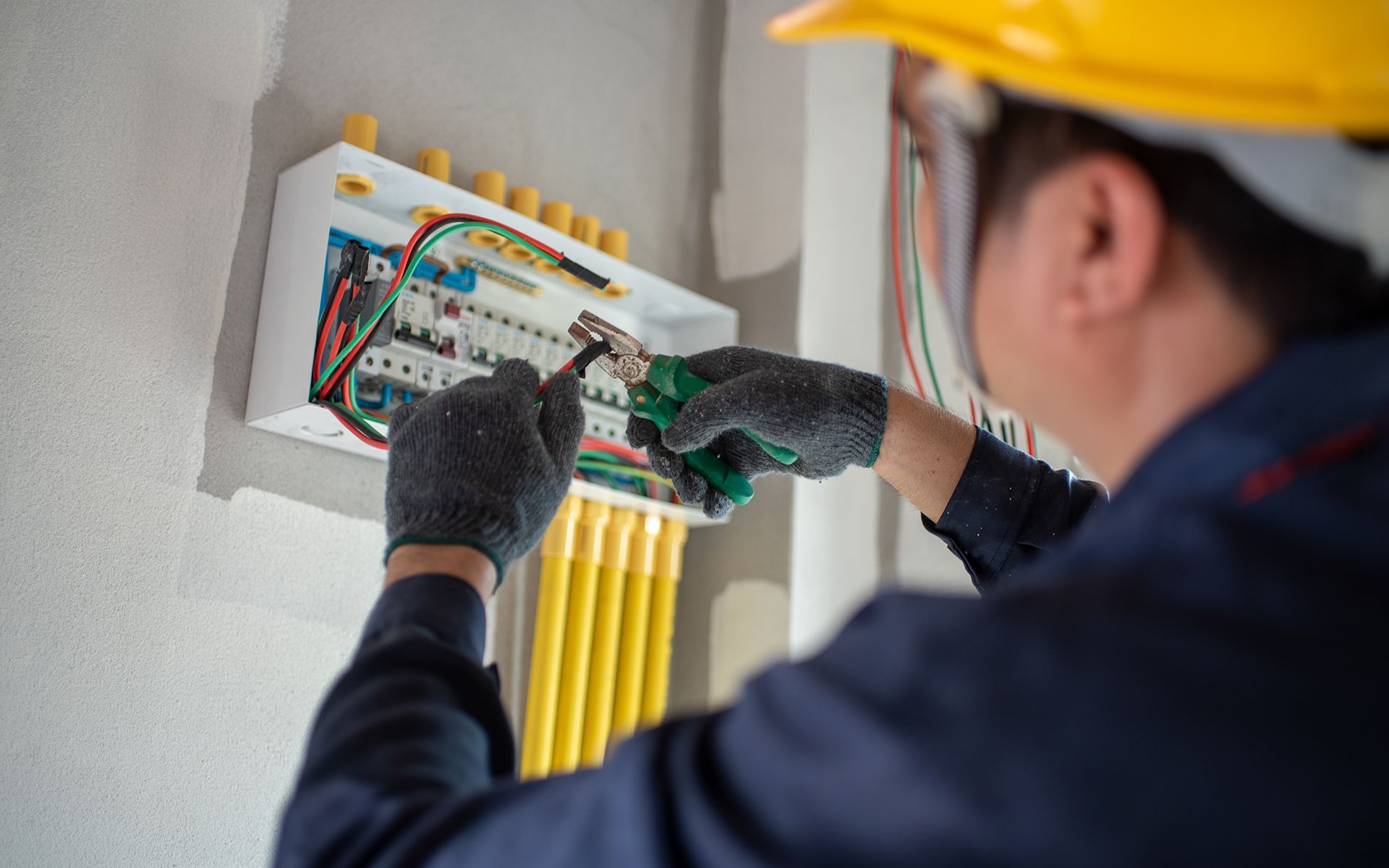 Electrician in gloves and hardhat working on electrical panel.