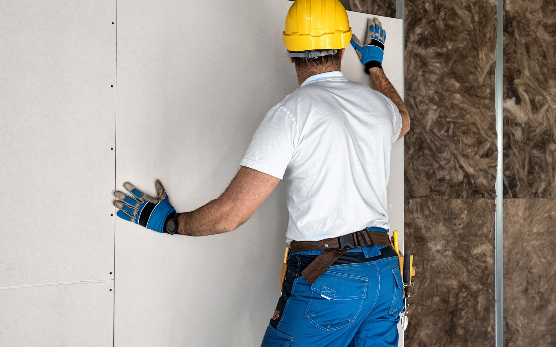Construction worker installing drywall, wearing gloves and a hard hat.
