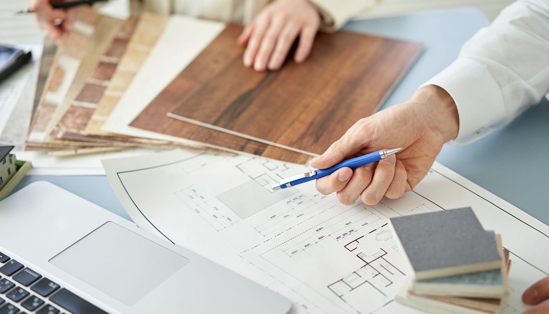 Hands pointing at blueprints, surrounded by flooring samples and laptop.