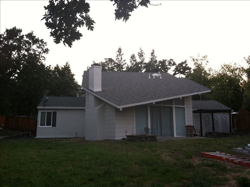 Gray house with slanted roof, chimney, windows, and small shed, surrounded by trees and a grassy yard.