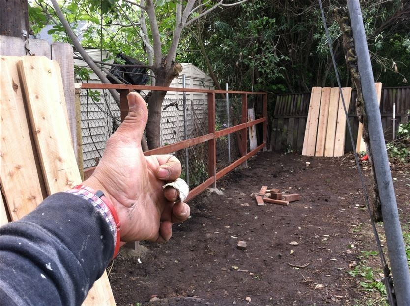 A hand with a thumbs up gesture in front of a partially built brown metal fence in a yard with wooden planks.