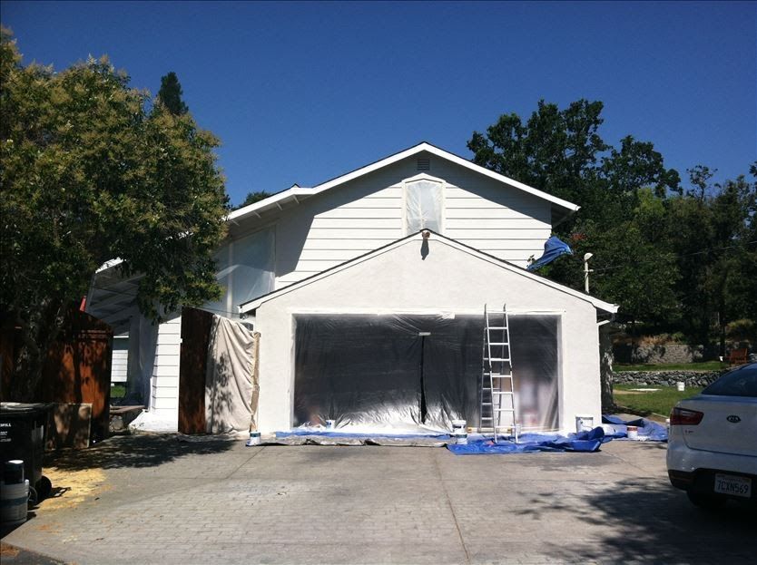 House exterior during painting, garage door covered, ladder present, blue tarp on driveway.