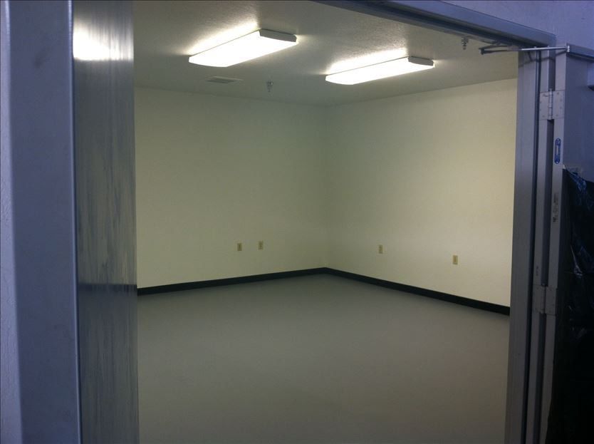 Empty, white-walled room with gray floor, viewed from doorway. Fluorescent lights illuminate the space.