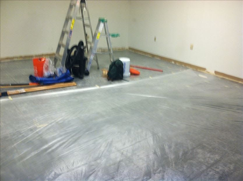 Room with floor covered in plastic sheeting, ladders, and painting supplies. Walls are white.
