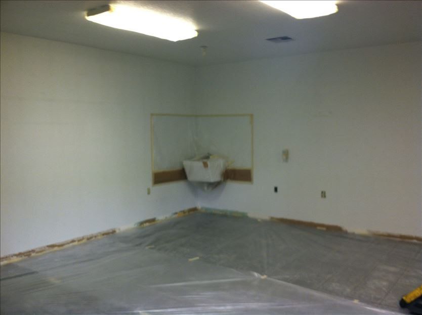 Empty room with white walls, fluorescent lights, and a corner sink. Gray floor with exposed edges.