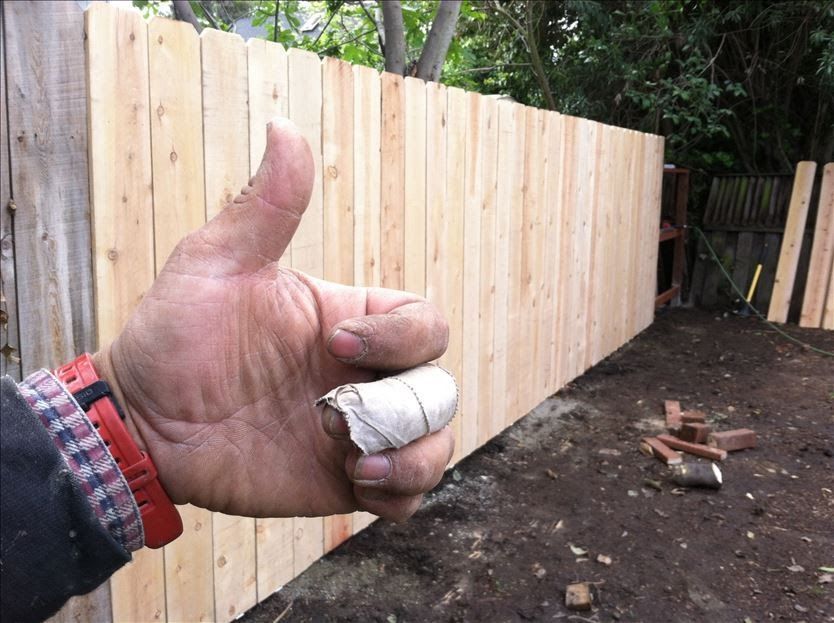 Person's hand with bandaged finger giving thumbs up by a newly built wooden fence.