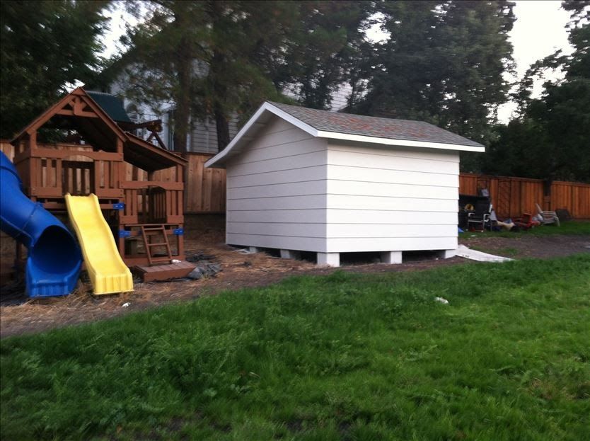 White shed with a gray roof next to a wooden playset in a grassy backyard, brown fence in the background.
