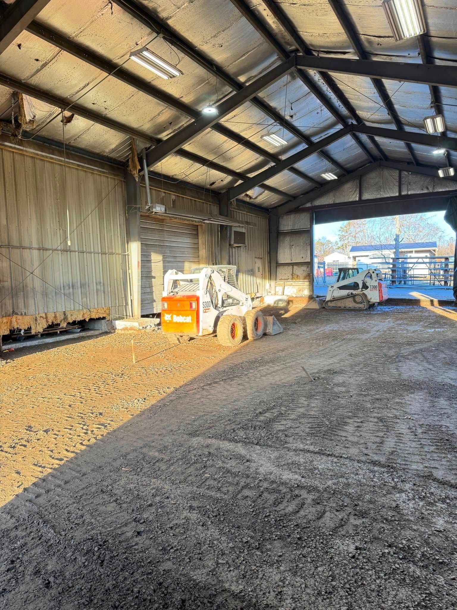 Inside a construction site: A Bobcat tractor works on a gravel ground under a metal roof.