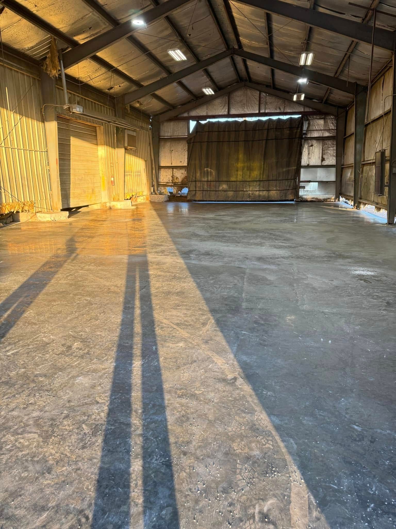 Inside a large, empty metal warehouse with concrete floor, sunlit shadows stretch across the floor.