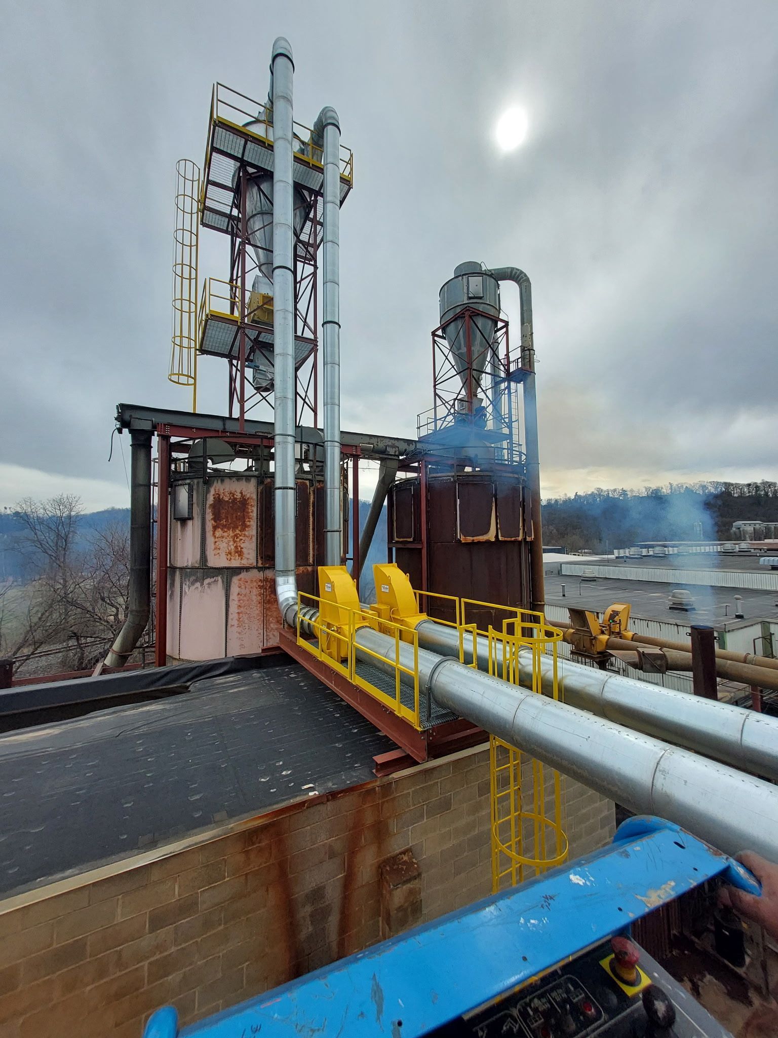 Industrial facility with tall metal structures, pipes, and smoke against a cloudy sky.