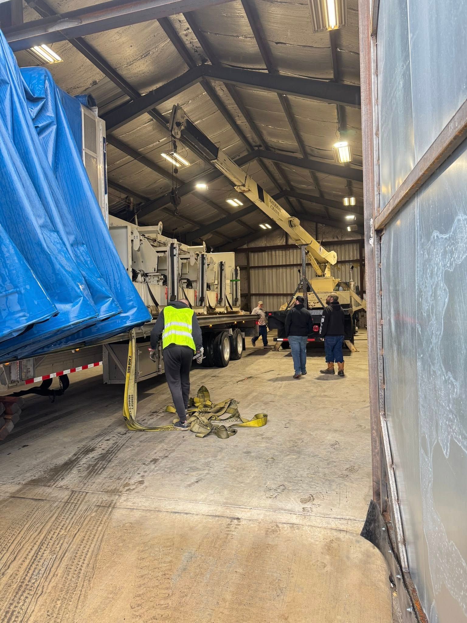 Workers unload a large blue tarp from a truck inside a warehouse using a crane.