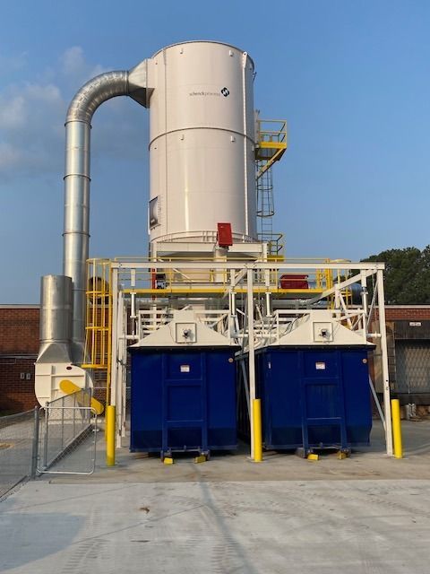 Dust collection system with white silo, blue bins, and metal piping. Outdoors, against a blue sky.
