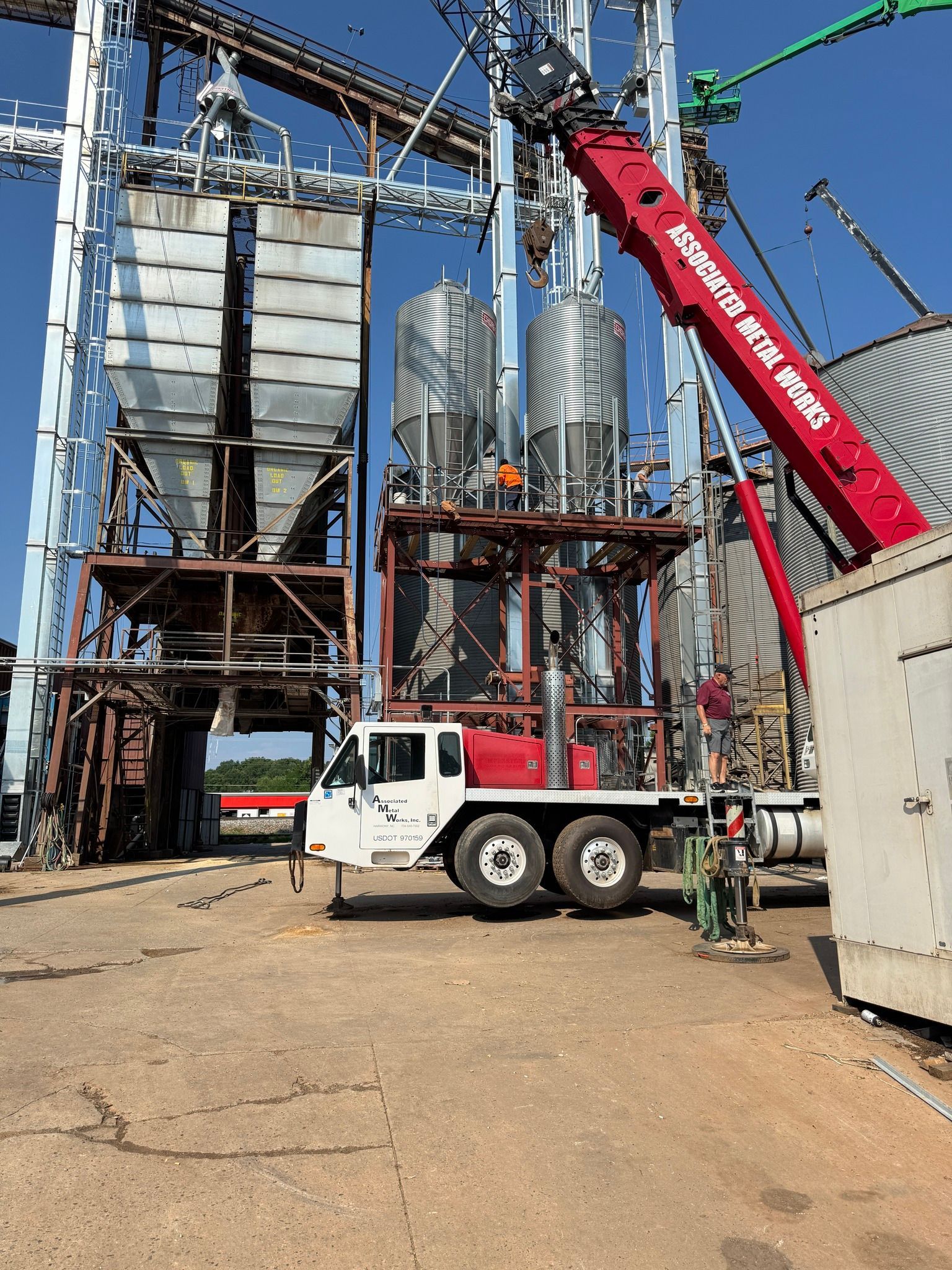 A red crane lifts a structure at a grain elevator with a white truck supporting it on a sunny day.
