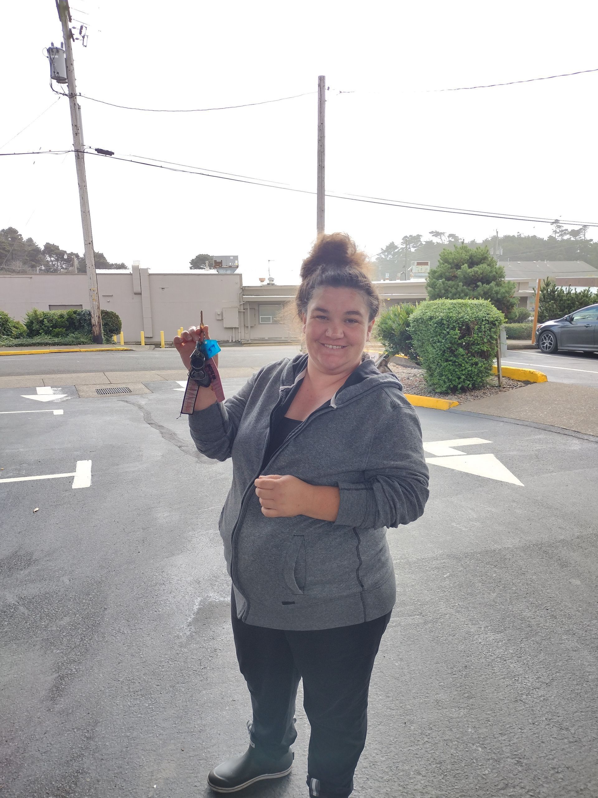 Woman in gray hoodie holding keys in a parking lot, smiling with cloudy sky background.