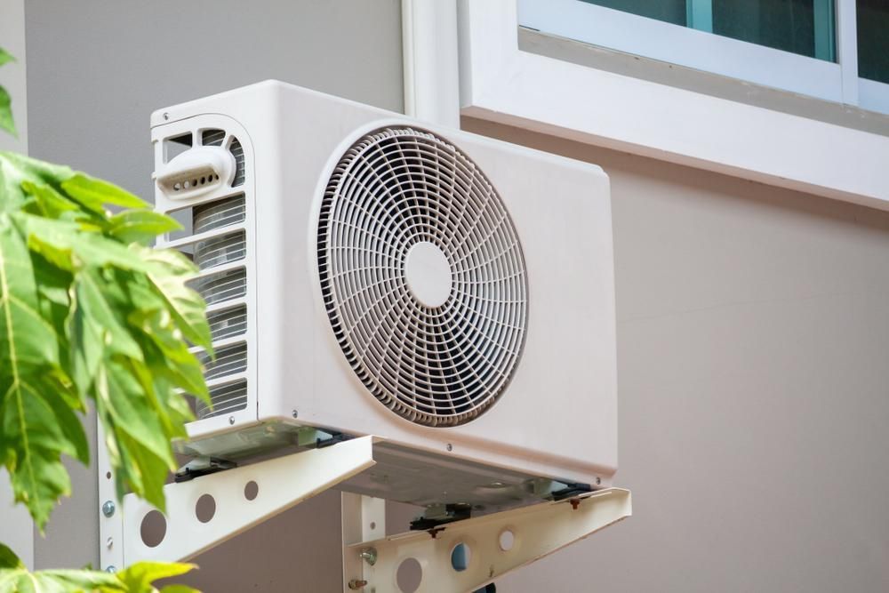 A White Air Conditioner Is Mounted On The Side Of A Building Next To A Window — Jervis Bay Airconditioning In Sussex Inlet, NSW