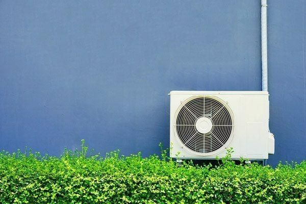 A White Air Conditioner Is Mounted To A Blue Wall Next To A Hedge — Jervis Bay Airconditioning In Albion Park, NSW