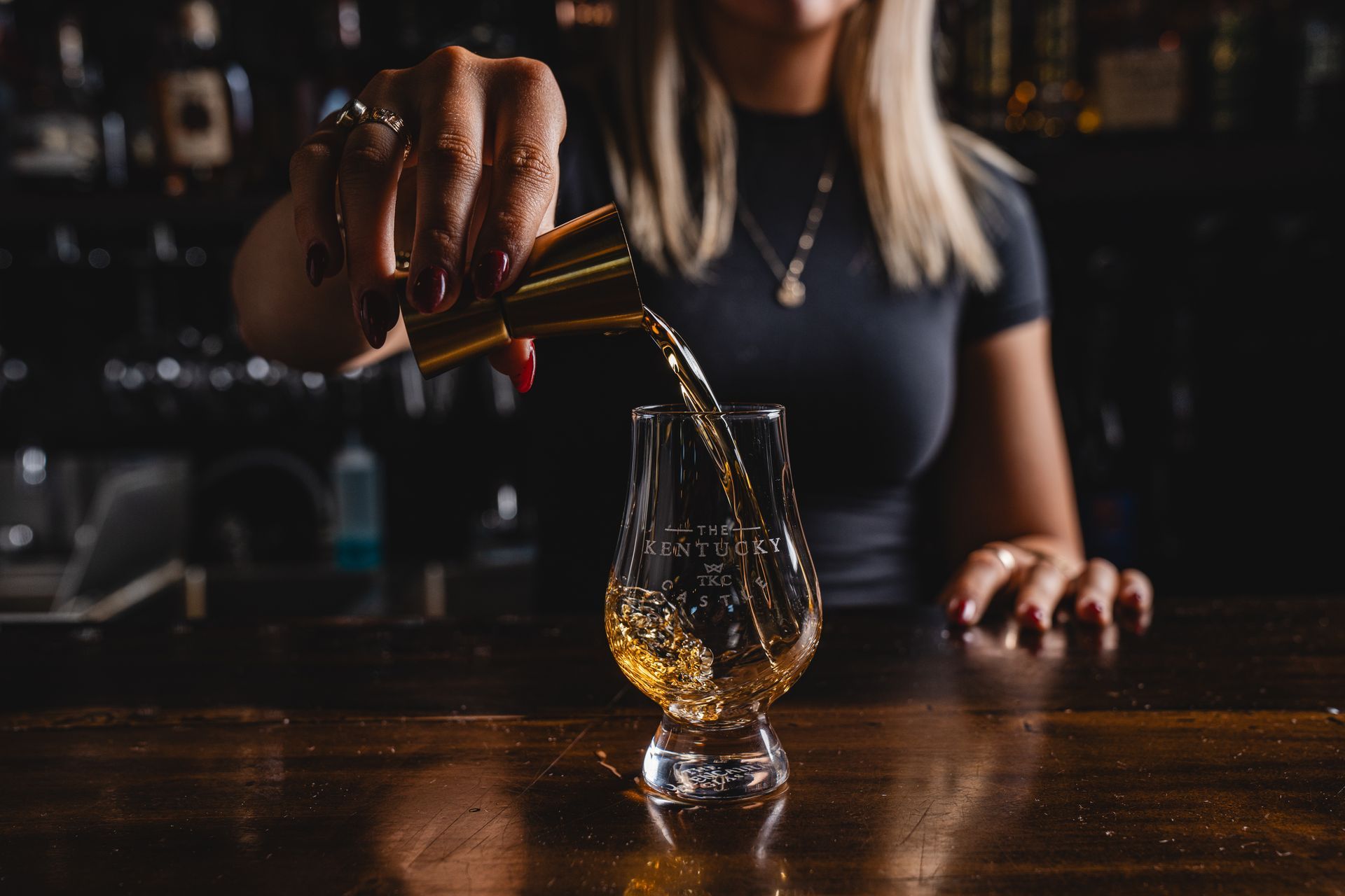 Bartender pouring amber liquid from a jigger into a tulip-shaped glass at a bar.