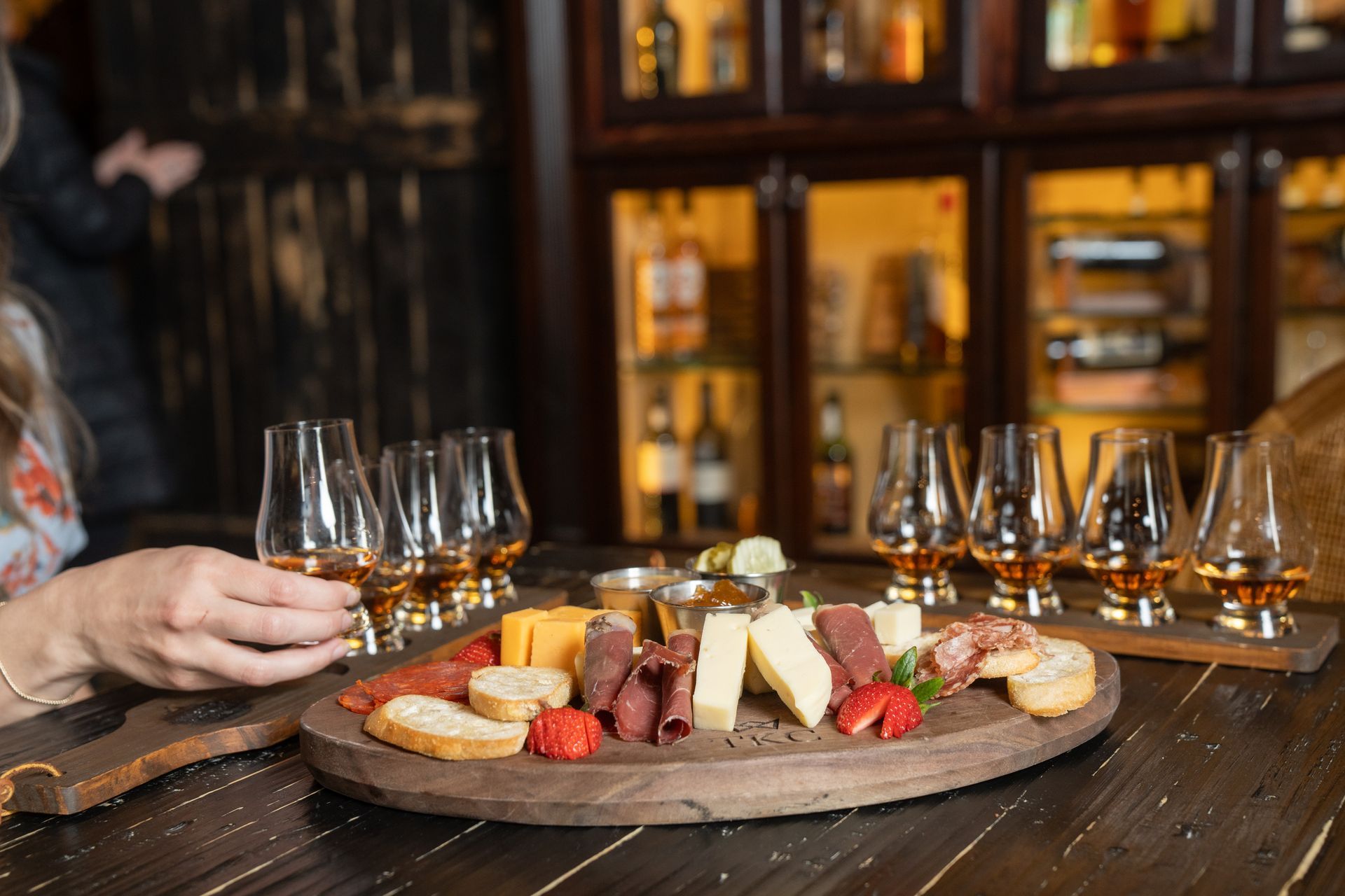 Woman holding a whiskey glass, charcuterie board with glasses of whiskey in a pub setting.