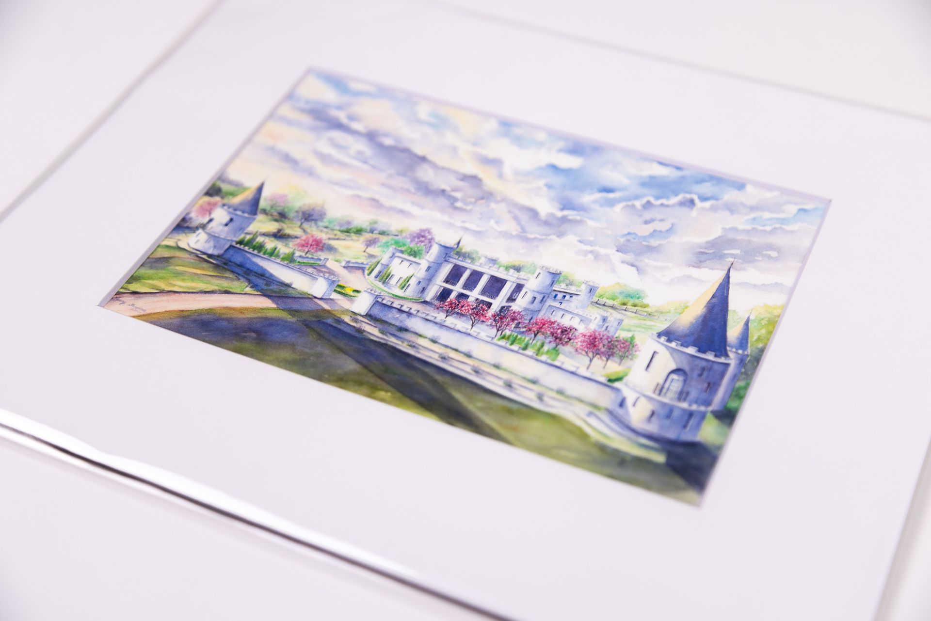 Watercolor painting of a white castle with towers, trees, and blue sky, mounted in a white mat.