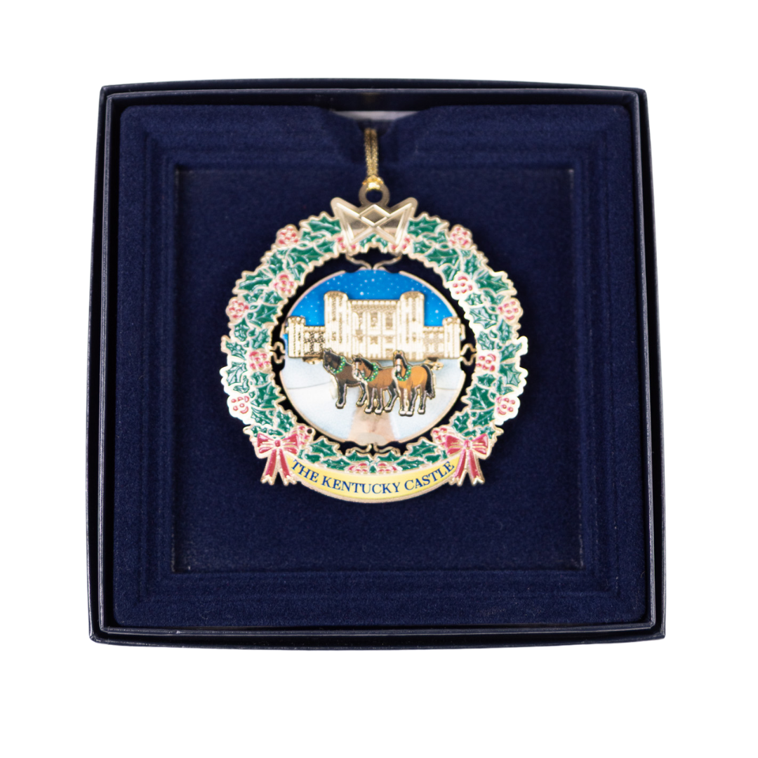 Ornament depicting a castle scene within a wreath, displayed in a blue box.