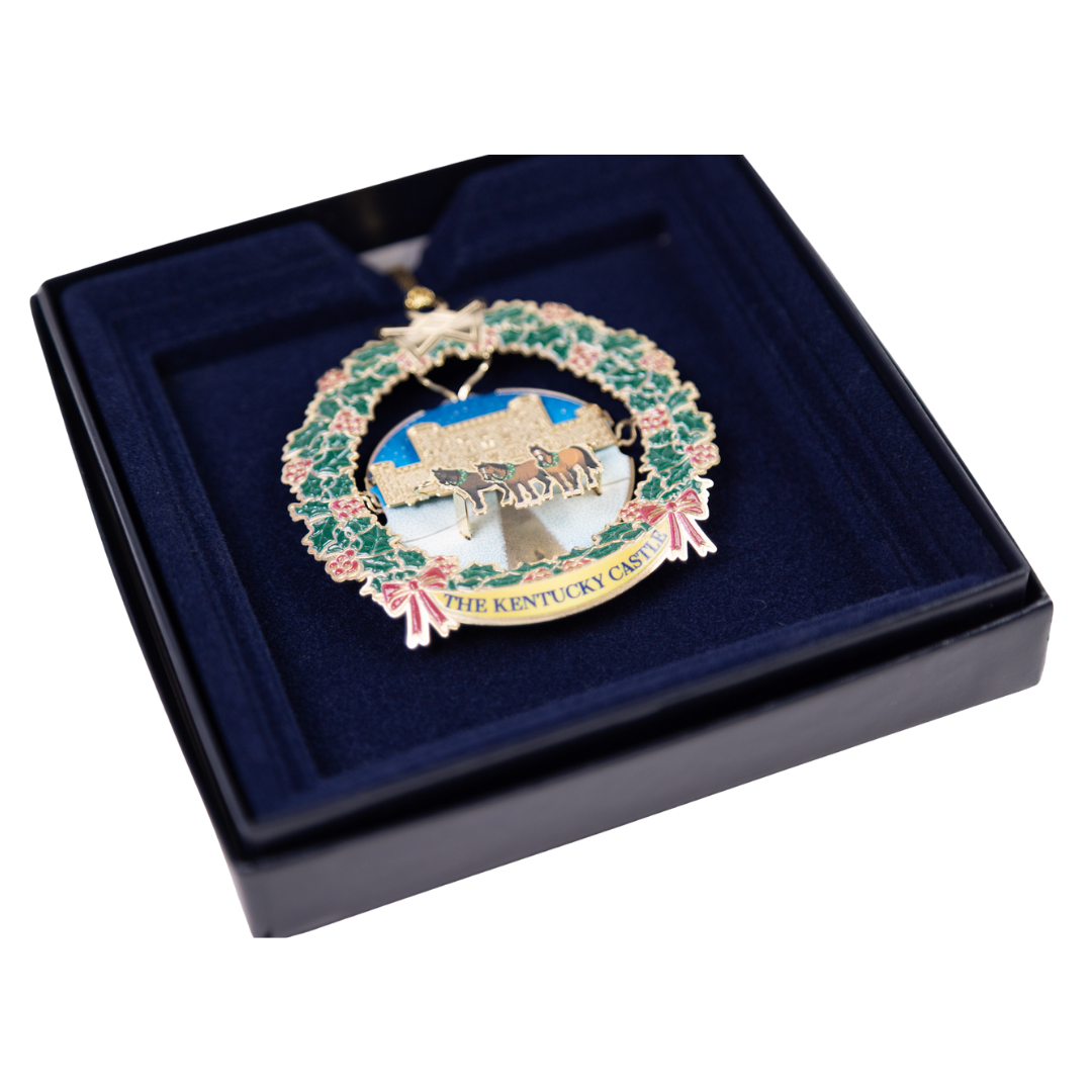 Ornament depicting a castle scene within a wreath, displayed in a blue box.