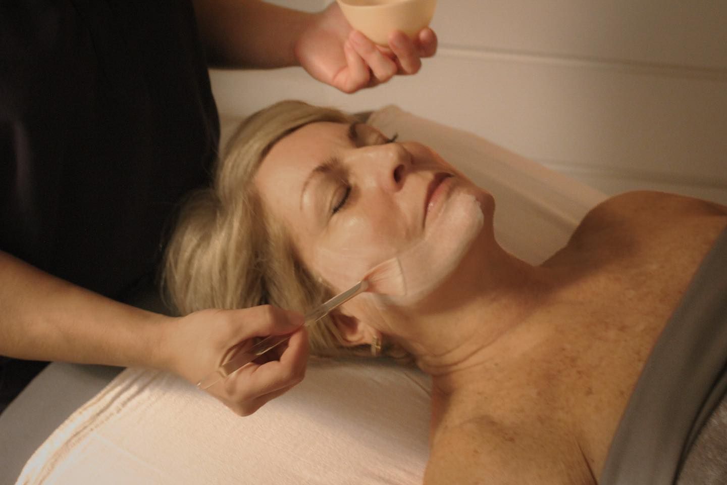 Woman receiving facial treatment; therapist applying a mask with a spatula.