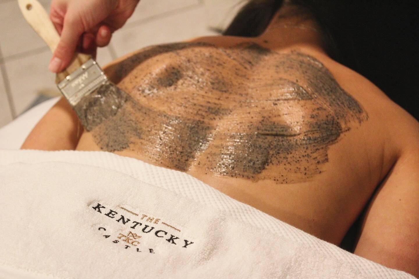 Person getting back scrub at a spa with brush applicator. Kentucky spa towel visible.