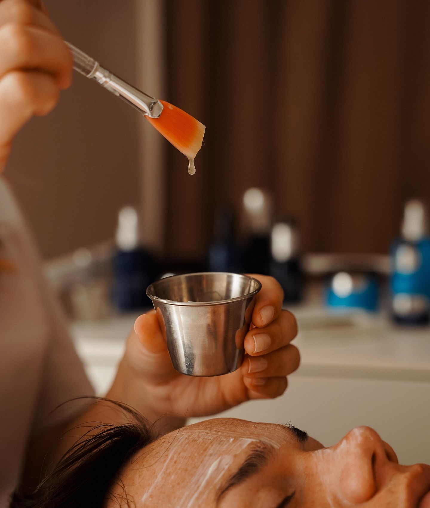 A cosmetician applies a facial treatment from a brush above a client's face.