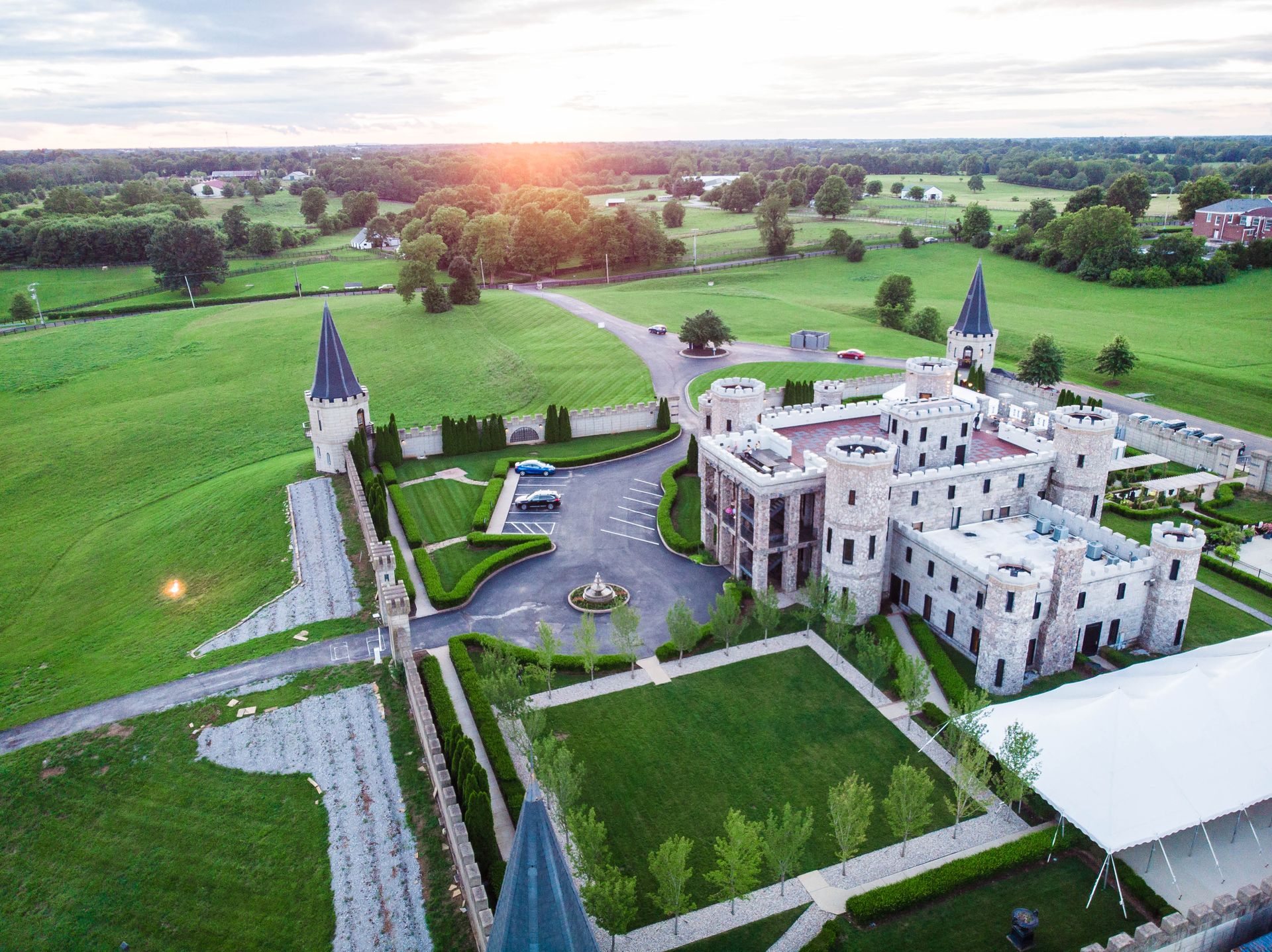 Castle with towers and a large lawn, surrounded by green fields, at sunset.