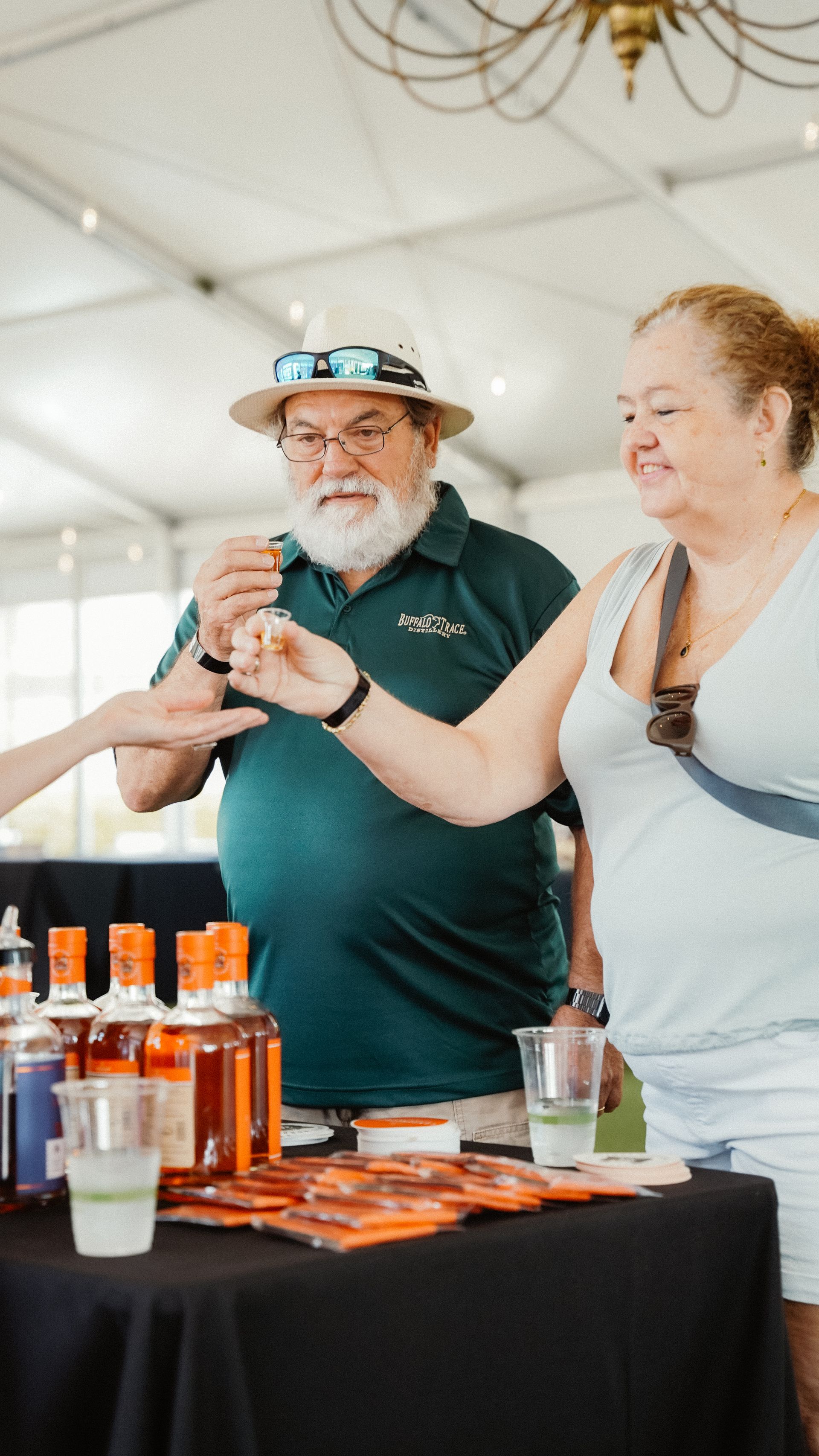 Couple tasting a drink at a vendor booth. Man in hat, woman smiling. Bottles and glasses on the table.
