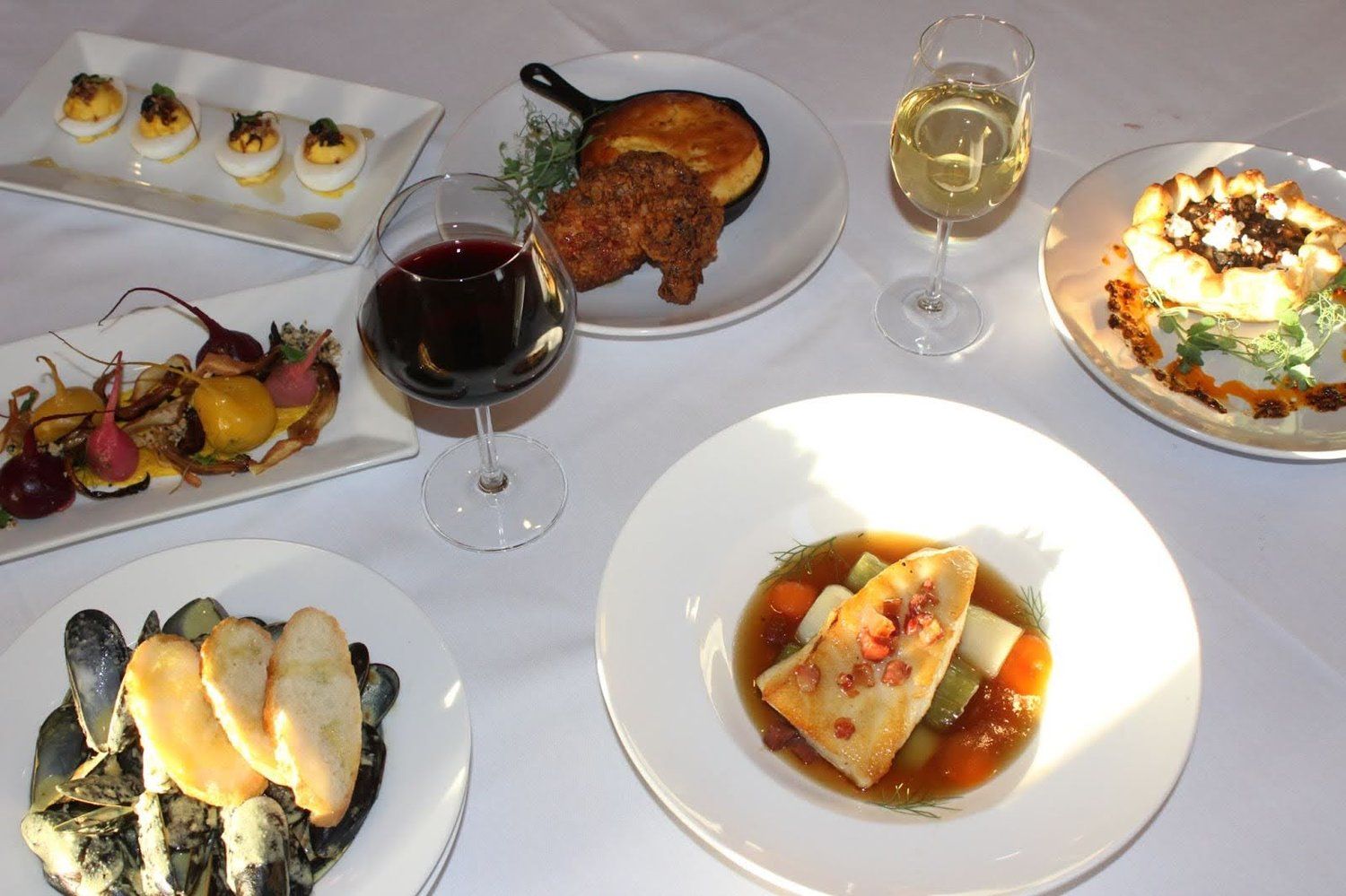 Fine dining table with several plated dishes, glasses of wine, and dessert on a white tablecloth